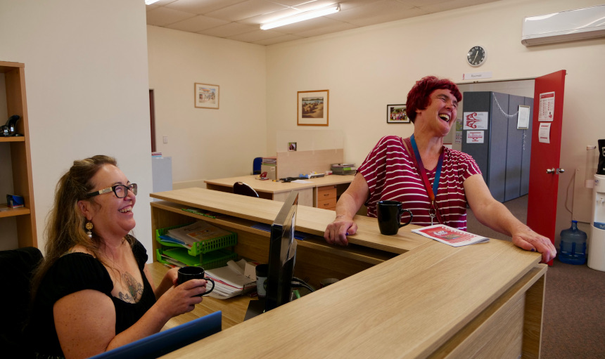 Sara (left) sitting behind the reception desk with Trish (right) leaning on the outside laughing