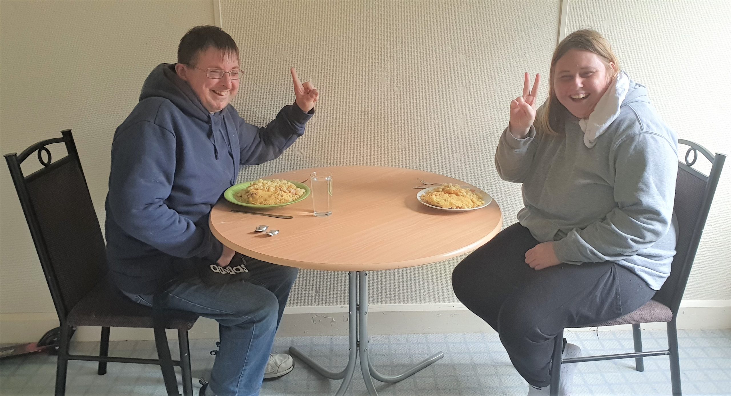 Jordan (left) and Courtney (right) sitting down at a table holding the peace sign up. They are both smiling with a pasta dish on the table.
